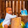 Tristan joined the competition — help win amazing prizes! toddler, child, barefoot, wooden_stump, balloons, cake, celebration, indoor, floor, wooden_fence, curious, standing, white_clothing, party, decor, baby, smiling, portrait, festive, colorful
