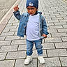 toddler, child, denim_jacket, blue_beanie, mickey_mouse, white_tshirt, jeans, white_sneakers, paved_street, outdoor, serious_expression, standing, urban, sidewalk, fashion, casual_clothing, person, young_child, cold_weather, street
