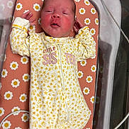 Tiana is registered to the contest to win money with this photo: newborn, baby, infant, child, portrait, lying_down, bassinet, crib, floral_pattern, onesie, yellow_clothing, white_clothing, pink_background, face, hands, indoor, nursery, sleeping, awake, hospital_bassinet