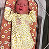 newborn, baby, infant, child, portrait, lying_down, bassinet, crib, floral_pattern, onesie, yellow_clothing, white_clothing, pink_background, face, hands, indoor, nursery, sleeping, awake, hospital_bassinet