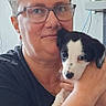 Baîka a rejoint le concours — aidez-le/la à gagner de superbes lots ! person, glasses, puppy, dog, indoor, holding, black_and_white, short_hair, smile, face, wall_pattern, sitting, casual_clothing, portrait, cute, pet, affection, young_dog, human, closeup