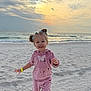 child, toddler, beach, sand, ocean, waves, sunset, sky, clouds, smile, pink_outfit, hoodie, sweatpants, crocs, hair_buns, wristband, footprints, horizon, playful, portrait
