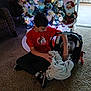 boy, child, baby, car_seat, blanket, christmas_tree, ornaments, lights, santa_decoration, carpet, indoor, holiday, home, red_shirt, sneakers, carpeted_floor, window, decorations, present, infant