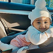 Amelia is registered to the contest to win money with this photo: baby, child, car_seat, hat, pom_poms, white_clothing, gloves, tights, pink_skirt, smile, face, indoors, car_interior, seatbelt, sunlight, warm_clothing, cozy, portrait, infant, cute
