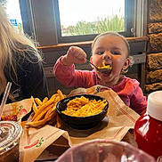 Cali joined the competition — help win amazing prizes! toddler, child, food, macaroni_and_cheese, french_fries, spoon, table, restaurant, window, drink, ketchup, person, pink_clothing, indoor, stone_wall, happy, eating, meal, cute, closeup