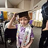 child, graduation_cap, sash, indoor, celebration, person, purple_shirt, seated_adult, table, chair, carpet, event, window, light, hair, face, people, room, casual, smile