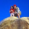 child, young_man, rock, outdoor, blue_sky, red_shirt, cap, hoodie, shorts, smile, nature, adventure, holding, happy, portrait, person, daylight, clothing, curly_hair, casual