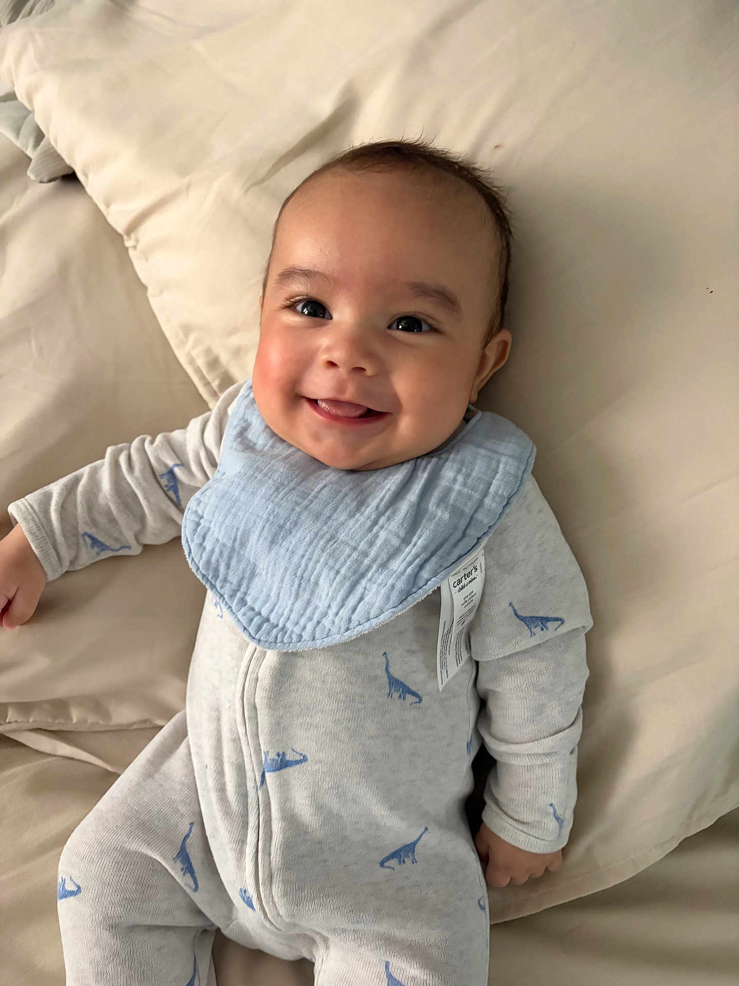 Emiliano joined the competition — help win amazing prizes! baby, smiling, infant, onesie, bib, dinosaur_pattern, bed, pillow, happy, child, cute, portrait, sleepwear, lying_down, indoor, face, skin, head, hand, young