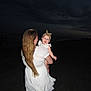 Harper is registered to the contest to win money with this photo: baby, woman, beach, ocean, night, smile, white_dress, headband, holding, child, outdoor, dark_sky, waves, sand, portrait, person, long_hair, happy, evening, family