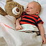 baby, teddy_bear, blanket, smile, bed, infant, plush_toy, striped_clothing, cute, sleeping_area, comfort, child, soft_toy, happy, resting, indoors, cozy, lying_down, skin, head