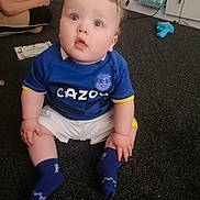 Kingston is registered to the contest to win money with this photo: baby, child, indoor, carpet, blue_jersey, sitting, curious, person, game_controller, toy, couch, sock, white_shorts, floor, hair, hand, face, eyes, mouth, furniture