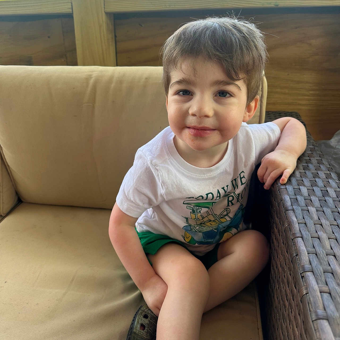 Kyler is registered to the contest to win money with this photo: child, boy, smile, white_tshirt, green_shorts, couch, beige_cushion, wicker_furniture, wooden_wall, outdoor, greenery, portrait, casual, happy, seated, short_hair, daylight, relaxed, cute, young
