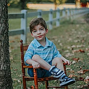 Kyler is registered to the contest to win money with this photo: child, boy, rocking_chair, outdoor, grass, tree, leaves, fence, blue_shirt, shorts, sneakers, smile, portrait, nature, fall, casual, sitting, daylight, cute, relaxed