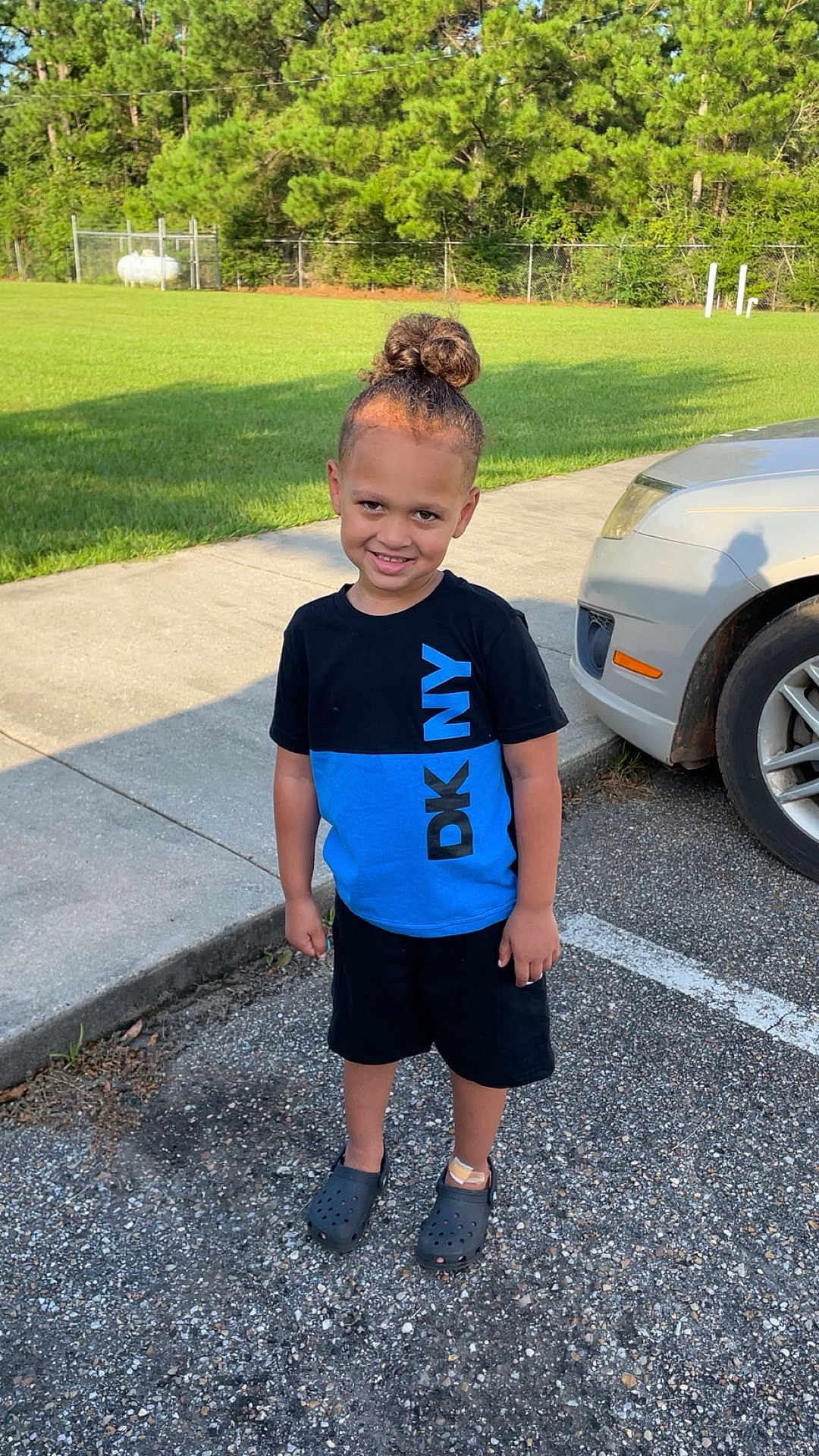 Jamarcus joined the competition — help win amazing prizes! car, casual_clothing, child, concrete, crocs, daytime, grass, hair_bun, happy, outdoor, parking_lot, person, shadow, shorts, sidewalk, smiling, standing, sunlight, trees, tshirt