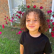 Jamarcus is registered to the contest to win money with this photo: black_tshirt, brick_wall, casual_clothing, child, curly_hair, cute, flower, garden, grass, greenery, happy, nature, outdoor, person, pink_flower, portrait, smile, sunlight, window, young
