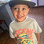 boy, cabinet, cap, cartoon, casual, child, clothing, countertop, floor, graphic_tshirt, happy, hat, indoors, kitchen, looking_up, person, smile, standing, tile_floor, tshirt