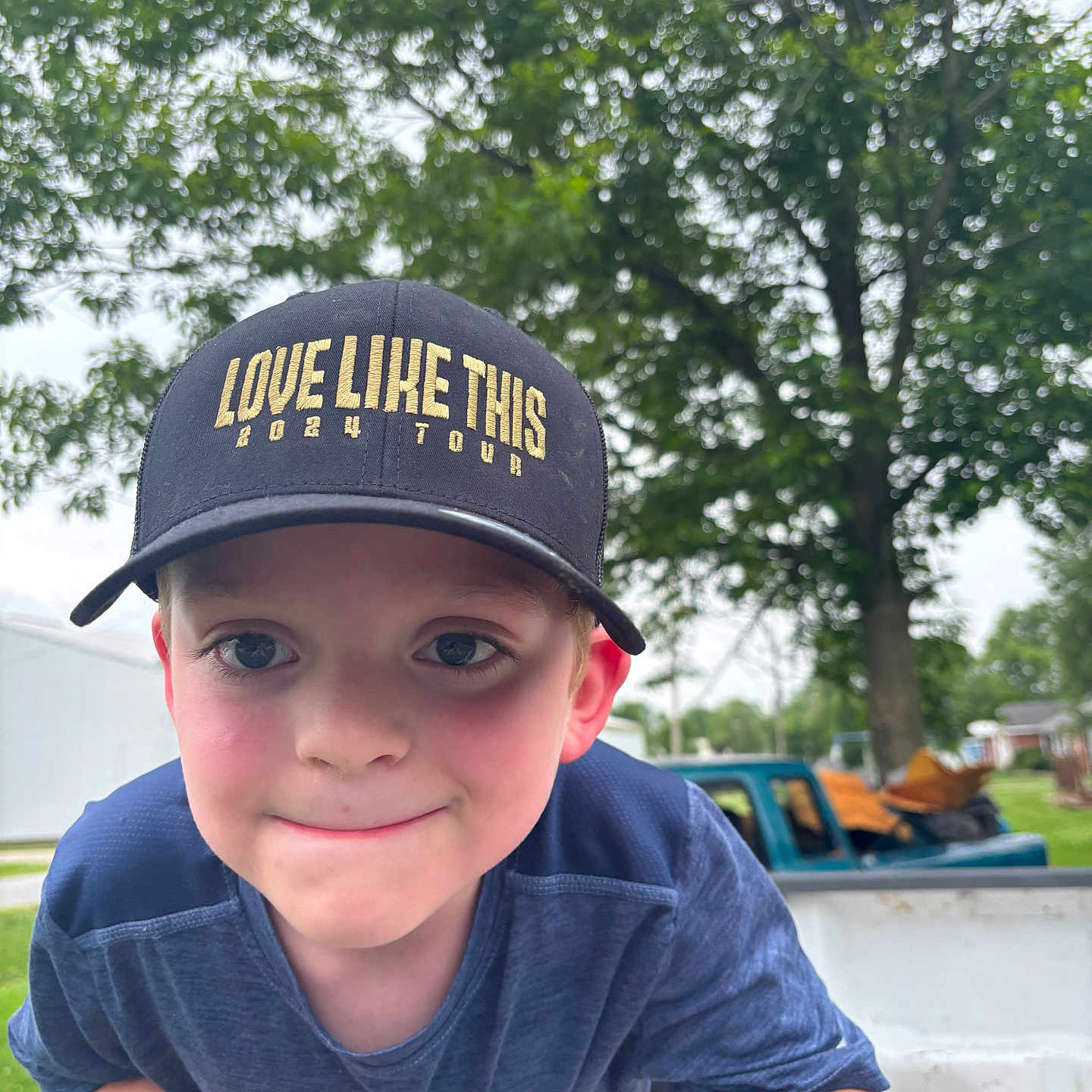 Grayson is registered to the contest to win money with this photo: blue, boy, cap, child, closeup, daylight, face, grass, greenery, hat, nature, outdoor, playful, portrait, shirt, short_sleeve, smile, tree, truck, vehicle