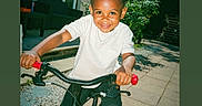Maylone participe au concours pour gagner de l'argent avec cette photo : child, bicycle, training_wheels, smiling, outdoor, sunny, house, trees, pavement, white_shirt, black_pants, black_shoes, red_handlebar, greenery, daylight, happy, person, play, fun, garden