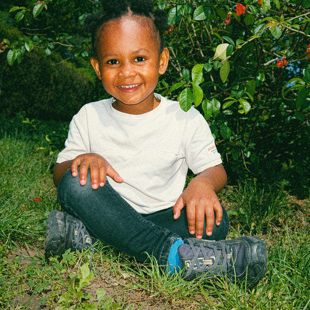 Maylone a rejoint le concours — aidez-le/la à gagner de superbes lots ! child, smiling, outdoor, grass, greenery, bush, nature, happy, person, casual_clothing, white_shirt, black_pants, shoes, sunlight, daylight, portrait, sitting, young_child, cute, playful