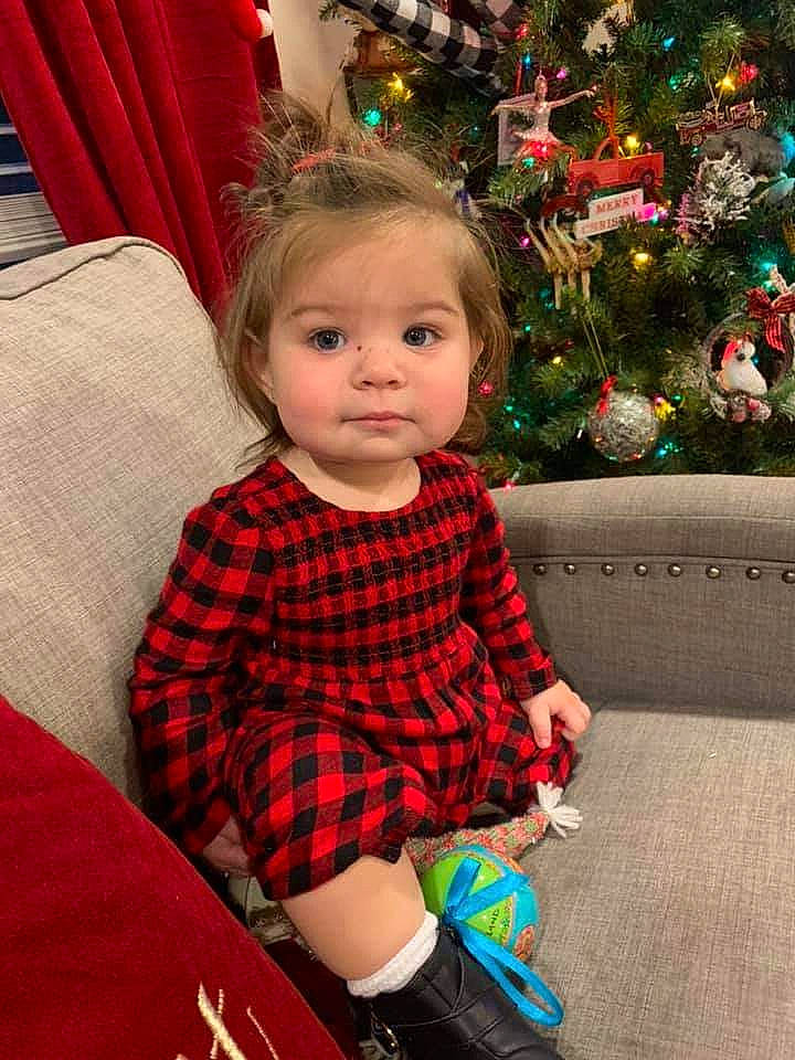 Eliana is registered to the contest to win money with this photo: baby_toddler_clothing, brown_hair, carmine, child, christmas_decoration, christmas_eve, christmas_ornament, christmas_tree, curtain, holiday, holiday_ornament, interior_design, nose, ornament, person, plaid, red, sitting, tartan, textile