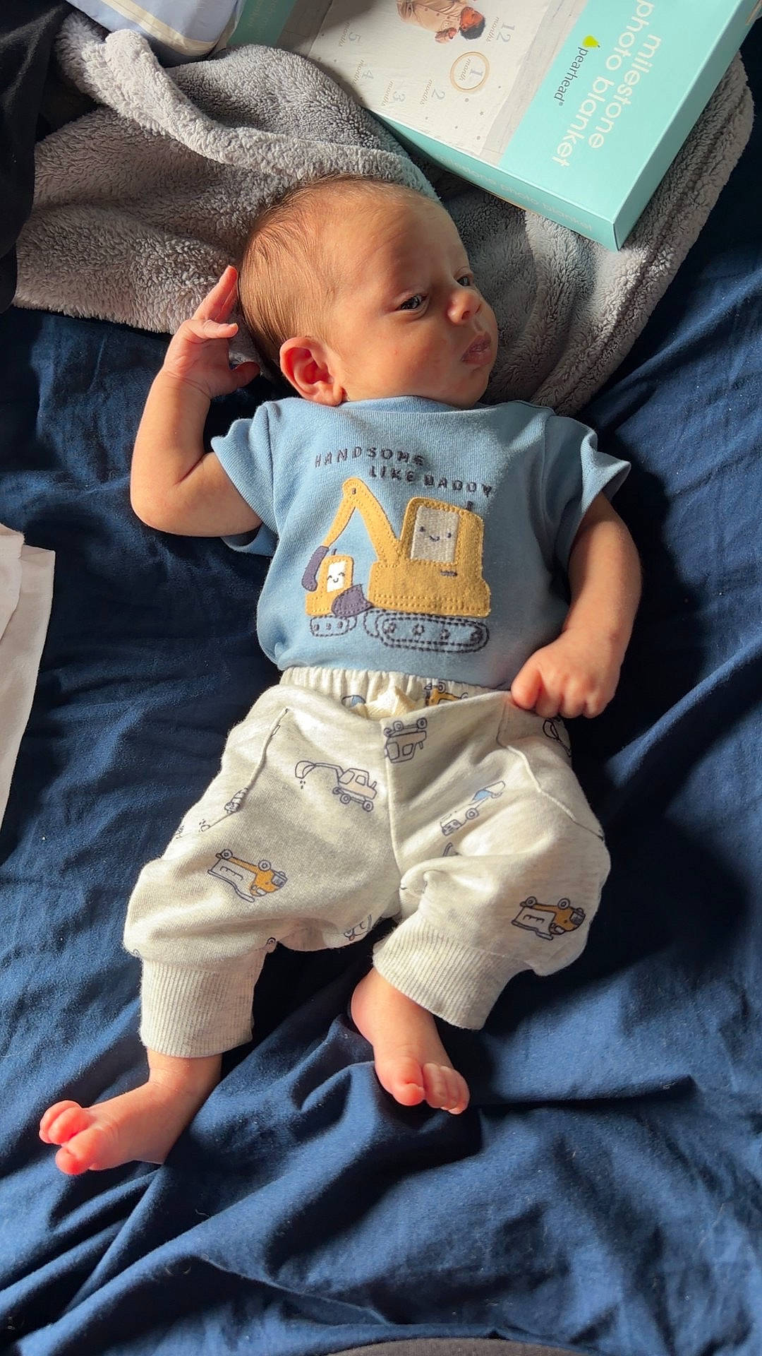 baby, bed, blanket, blue_shirt, casual, child, clothing, curious, cute_outfit, fabric, indoor, infant, lying_down, milestone_photo_blanket, newborn, portrait, relaxed, small_feet, soft_texture, toy_pattern