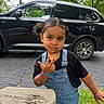 bench, black_shirt, black_suv, bracelet, car, child, curly_hair, daylight, denim_overalls, expression, girl, grass, hand_gesture, outdoor, park, pigtails, portrait, toddler, trees, wooden_bench