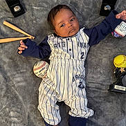 Andres is registered to the contest to win money with this photo: baby, infant, baseball_outfit, pinstripes, blanket, baseball, softball, mini_bats, rings, trophy, sports_memorabilia, cute, portrait, indoor, child, feet, hand, face, clothing, person