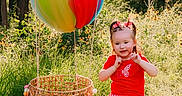 Blakely is registered to the contest to win money with this photo: child, girl, toddler, smiling, outdoor, forest, woodland, wildflowers, balloon, rainbow_balloon, basket, chair, red_shirt, checkered_shorts, hair_bows, shoes, sunlight, nature, portrait, playful