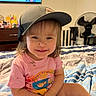 Blakely is registered to the contest to win money with this photo: bed, blanket, cap, cartoon, child, clothing, cute, dirt, dresser, fan, hair, happy, indoor, person, pink_shirt, room, sitting, smile, television, toddler