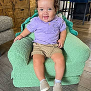 Samuel is registered to the contest to win money with this photo: toddler, child, smiling, curly_hair, purple_shirt, khaki_shorts, plush_chair, green_chair, shoes, socks, wooden_floor, wooden_cabinet, indoor, furniture, happy, person, portrait, living_room, cozy, cute
