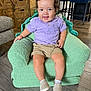 toddler, child, smiling, curly_hair, purple_shirt, khaki_shorts, plush_chair, green_chair, shoes, socks, wooden_floor, wooden_cabinet, indoor, furniture, happy, person, portrait, living_room, cozy, cute