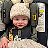 baby, child, car_seat, hat, pom_poms, blanket, pacifier, smile, face, hand, clothing, green, black, yellow, indoor, infant, cute, comfort, seatbelt, portrait