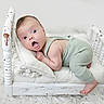 baby, infant, green_romper, small_bed, white_bed, wooden_bed, fluffy_rug, surprised_expression, wide_eyes, open_mouth, cushion, knitted_blanket, indoor, portrait, cute, child, resting, soft_lighting, white_background, cozy