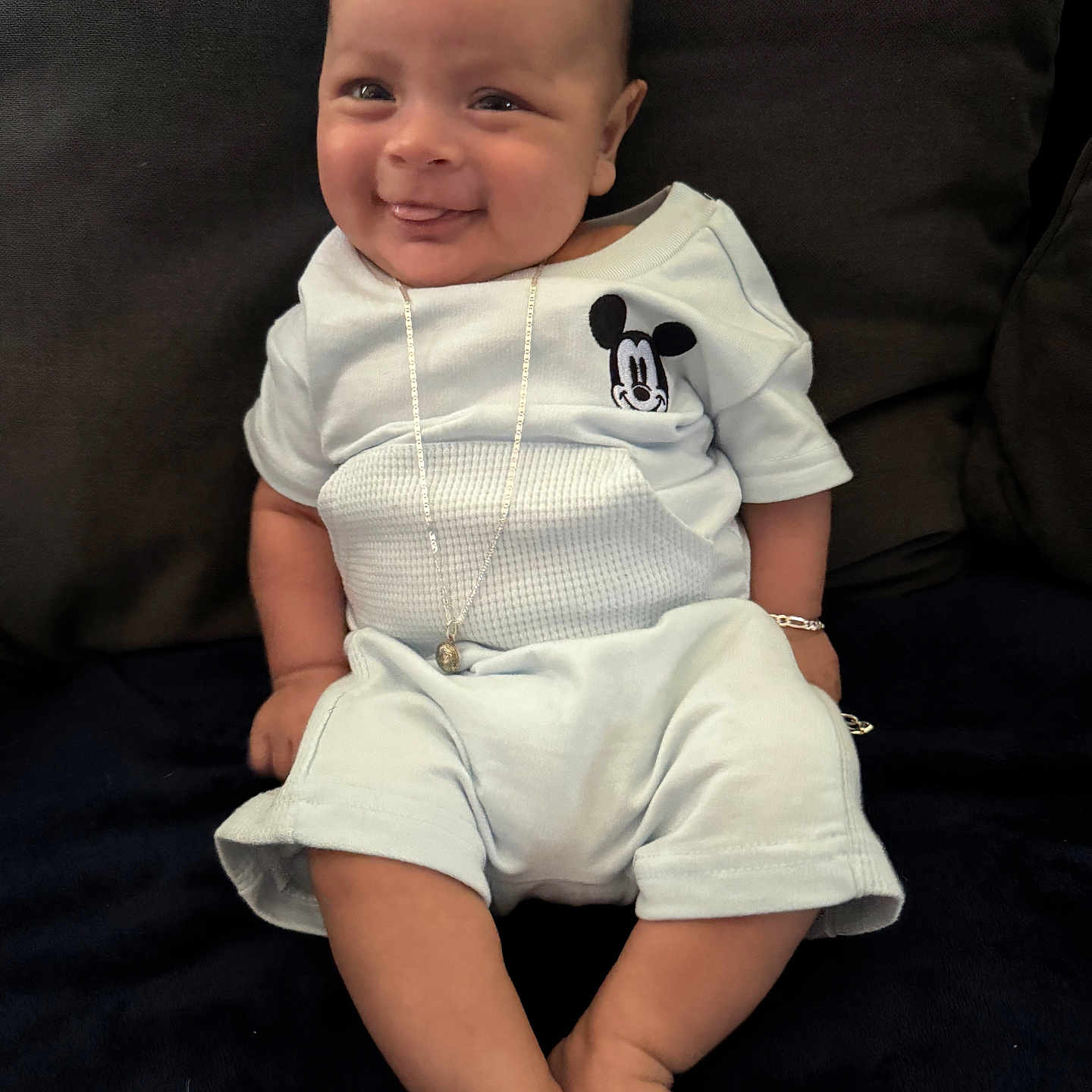 Frankko is registered to the contest to win money with this photo: baby, blue_outfit, bracelet, casual_clothing, child, comfortable, couch, cute, dark_background, happy, indoors, infant, mickey_mouse, necklace, person, plush, portrait, sitting, smiling, tongue_out
