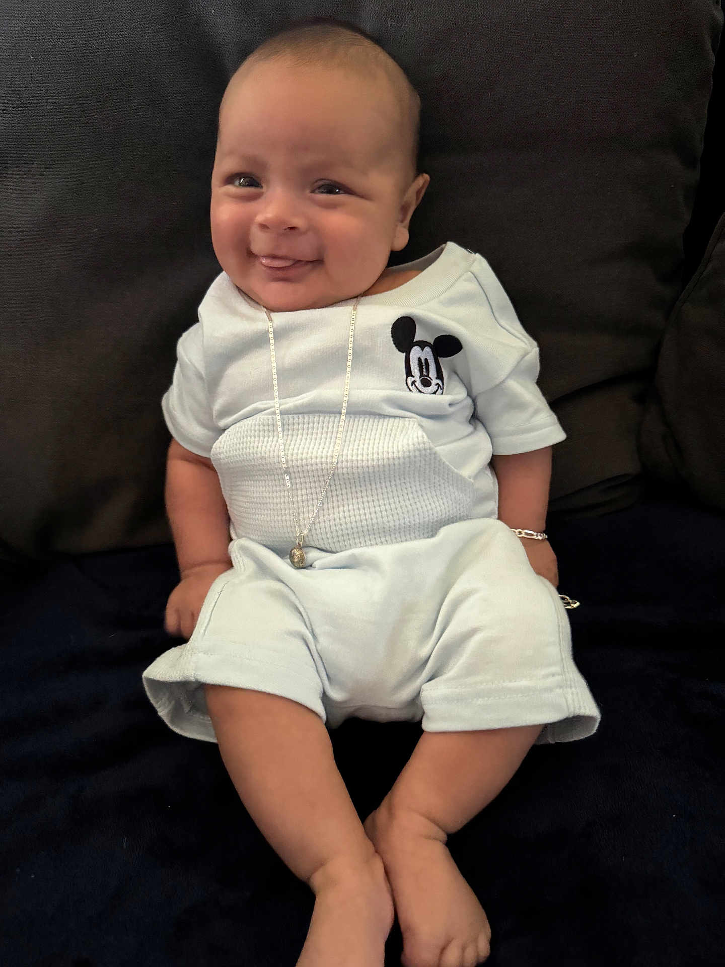 Frankko is registered to the contest to win money with this photo: baby, smiling, tongue_out, mickey_mouse, blue_outfit, necklace, bracelet, couch, dark_background, sitting, happy, infant, cute, indoors, person, child, comfortable, plush, casual_clothing, portrait