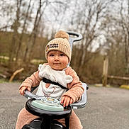Stiles a rejoint le concours — aidez-le/la à gagner de superbes lots ! child, toddler, baby, beanie, pompom, hat, ride_on_toy, toy_car, steering_wheel, bib, cozy_clothing, brown_clothing, outdoor, trees, blurred_background, pavement, hands, face, smile, portrait
