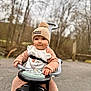 child, toddler, baby, beanie, pompom, hat, ride_on_toy, toy_car, steering_wheel, bib, cozy_clothing, brown_clothing, outdoor, trees, blurred_background, pavement, hands, face, smile, portrait