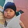baby, child, face, beanie, black_hat, baby_carrier, hand, outdoor, pavement, drain, clothing, cute, expressionless, person, young_child, closeup, infant, headwear, soft_texture, skin