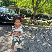 Alex is registered to the contest to win money with this photo: toddler, child, smiling, curly_hair, striped_shirt, green_shorts, white_shoes, asphalt, tree, shadows, parked_cars, sunny_day, outdoor, sidewalk, parking_lot, nature, happy, walking, daylight, casual_clothing