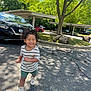 toddler, child, smiling, curly_hair, striped_shirt, green_shorts, white_shoes, asphalt, tree, shadows, parked_cars, sunny_day, outdoor, sidewalk, parking_lot, nature, happy, walking, daylight, casual_clothing