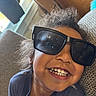 child, smile, sunglasses, curly_hair, indoor, couch, happy, face, person, casual_clothing, closeup, teeth, portrait, relaxing, cozy, window, light, hair, eyewear, fun