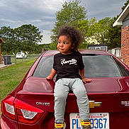 Malik is registered to the contest to win money with this photo: child, car, chevrolet, cruze, license_plate, tshirt, sneakers, grass, tree, cloudy_sky, brick_house, fence, outdoor, person, vehicle, curly_hair, casual_clothing, daylight, side_view, sitting