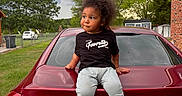 Malik is registered to the contest to win money with this photo: child, car, chevrolet, cruze, license_plate, tshirt, sneakers, grass, tree, cloudy_sky, brick_house, fence, outdoor, person, vehicle, curly_hair, casual_clothing, daylight, side_view, sitting