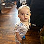 toddler, child, indoor, wooden_floor, pajamas, blue_shoes, curious, standing, person, face, young_child, hair_tied, footwear, home, blurred_background, furniture, expression, looking_up, casual_clothing, cute