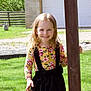 child, girl, smiling, outdoor, green_grass, wooden_post, flower_print, long_hair, sunlight, fence, garage, casual_clothing, happy, nature, daylight, person, standing, portrait, young_child, playful