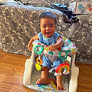 Clyde is registered to the contest to win money with this photo: baby, child, infant, smiling, sitting, activity_seat, baby_toys, colorful_toys, playtime, indoor, living_room, couch, patterned_fabric, wooden_floor, portrait, happy, blue_clothing, bare_feet, family_photo, home
