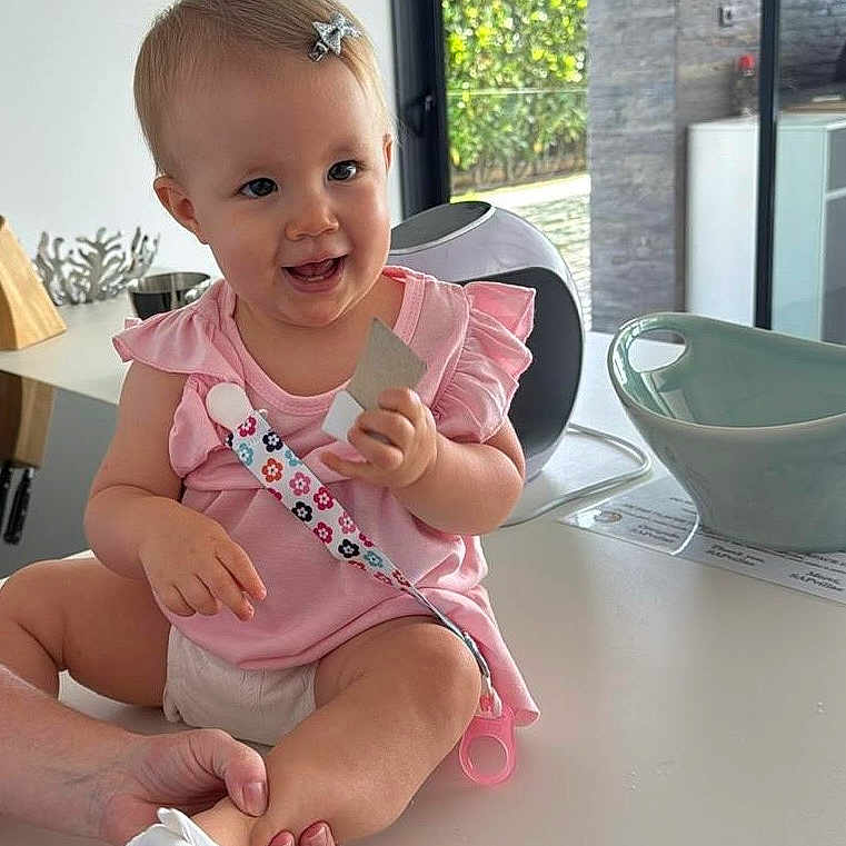 Mia participe au concours pour gagner de l'argent avec cette photo : baby, child, countertop, daylight, female, footwear, greenery, hair_clip, hand, happy, holding, indoor, kitchen, modern_interior, object, person, pink_clothing, sitting, smiling, window