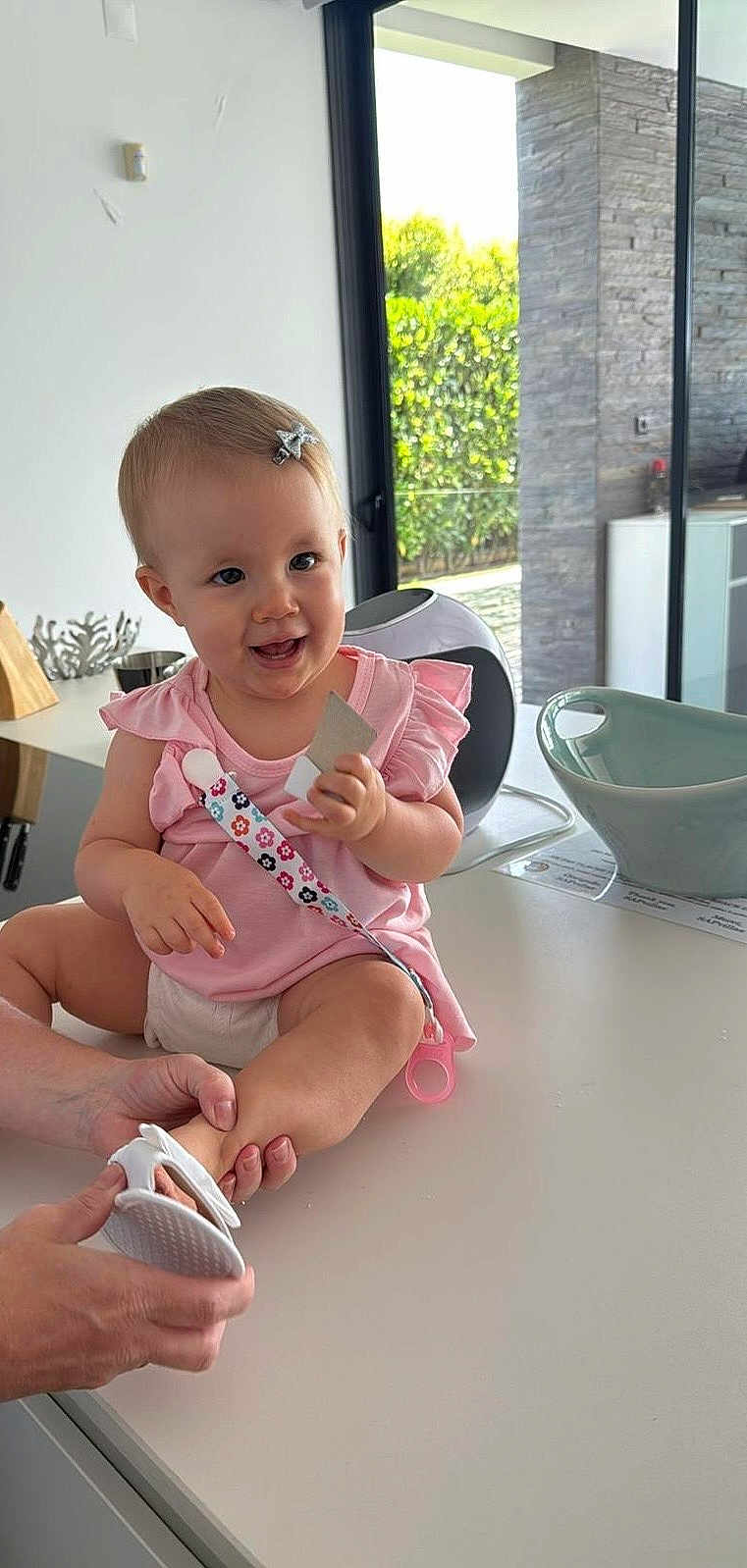 Mia participe au concours pour gagner de l'argent avec cette photo : baby, child, pink_clothing, smiling, indoor, kitchen, countertop, footwear, hand, modern_interior, window, greenery, happy, sitting, holding, object, hair_clip, female, person, daylight