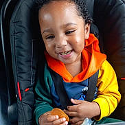 Ky-Mani a rejoint le concours — aidez-le/la à gagner de superbes lots ! toddler, child, car_seat, smiling, curly_hair, hoodie, orange_hood, yellow_sleeve, green_sleeve, snack, seatbelt, happy, baby, portrait, indoor, hands, face, cute, young_child, casual_clothing