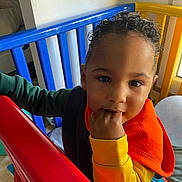 Ky-Mani participe au concours pour gagner de l'argent avec cette photo : toddler, child, curly_hair, teething, playpen, colorful, red, blue, yellow, fingers_in_mouth, indoor, toy, window, natural_light, clothing, orange, yellow_sleeve, green_sleeve, face, person